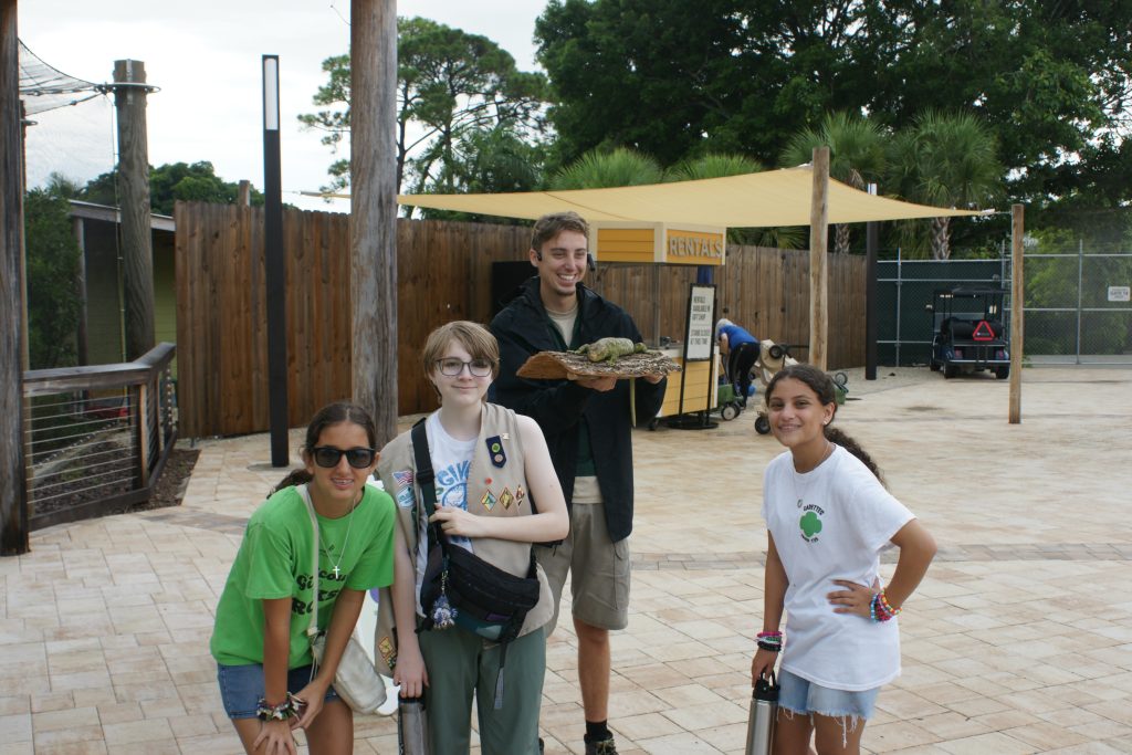 3 girl scouts standing in front of a animal trainer. the trainer is holding a piece of wood that has a green and black prehensile-tailed skink on it.